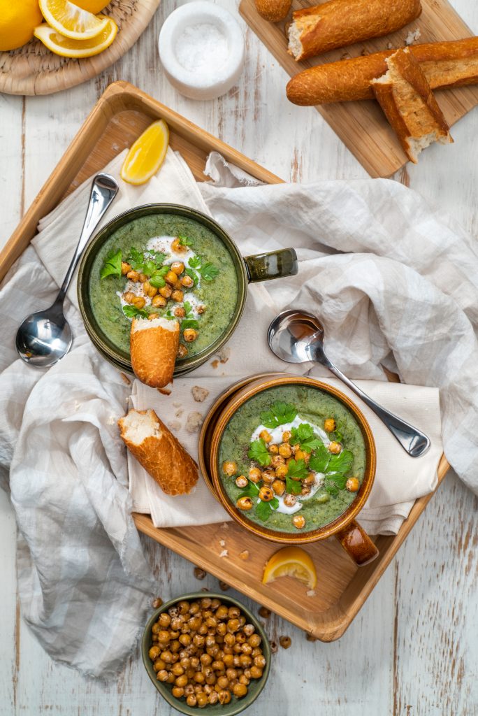 Two bowls of broccoli and potato soup topped with chickpeas and cream and served with baguette and lemon wedges on a wooden tray