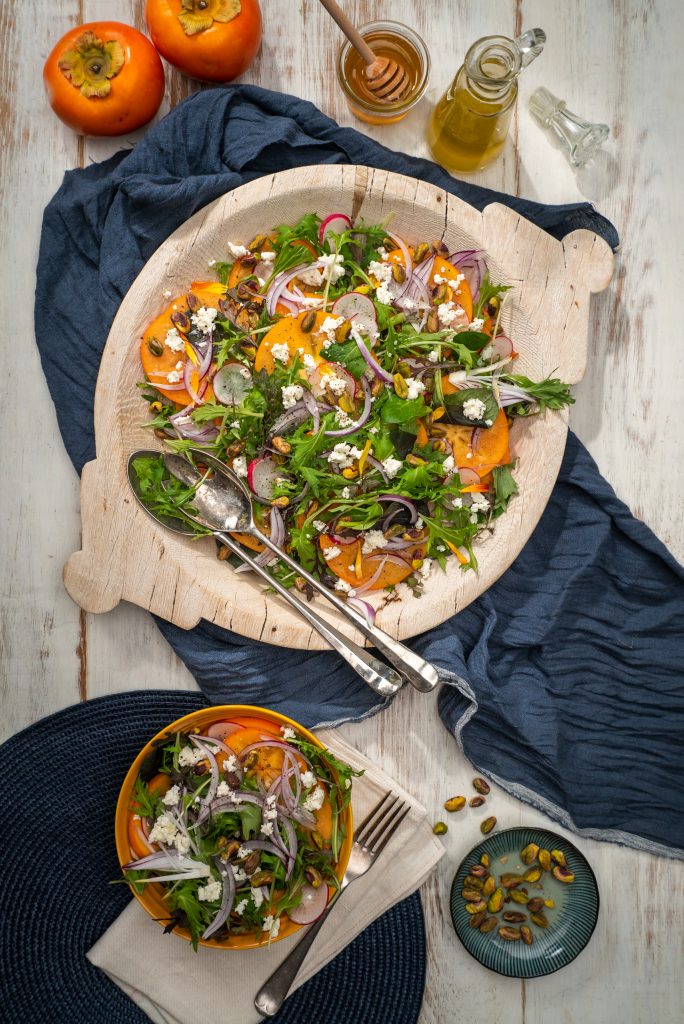 Persimmon, salad greens, red onion and feta salad in a wooden bowl with serving spoons on a blue cloth