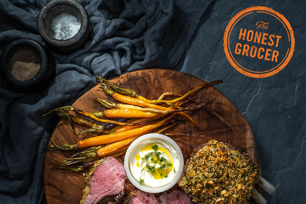 Crusted lamb rack with baby carrots and dips on a wooden board with The Honest Grocer logo