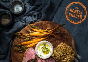 Crusted lamb rack with baby carrots and dips on a wooden board with The Honest Grocer logo